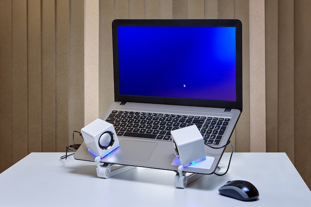 테이블 표면에는 냉각 홀더에 파란색 화면이 장착 된 노트북과 파란색 백라이트가있는 흰색 USB 스피커 2 개와 컴퓨터 마우스가 있습니다.