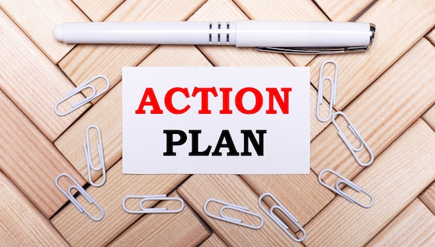 On a surface of wooden blocks, a white pen, white paper clips and a white card with the text ACTION PLAN. View from above.