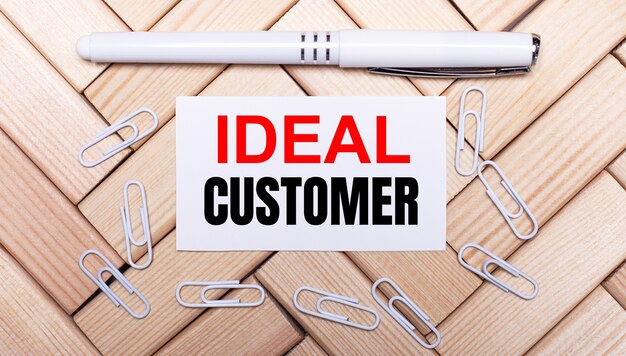 On a surface of wooden blocks, a white pen, white paper clips and a white card with the text IDEAL CUSTOMER