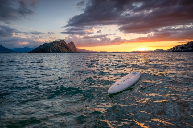 A surfboard lies in the water in a beautiful landscape while the sun sets leaving a red sky behind