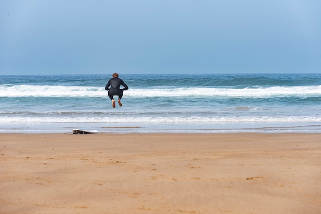 Surfer die op het strand naast zijn raad met het overzees op de achtergrond springt