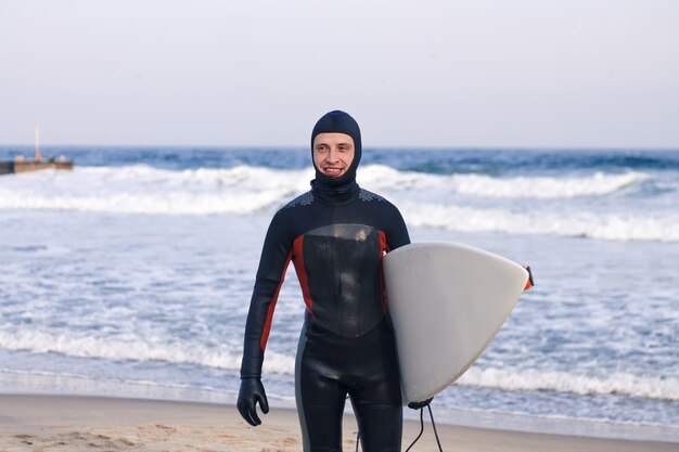 Surfer gaat uit het water met een wetsuit. Hij houdt een wit sufrboard vast