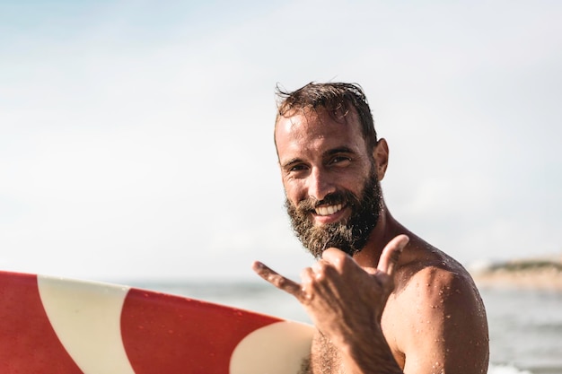 Surfer happy with surf surfing smiling doing hawaiian shaka\
hand sign for fun during surf session in ocean waves on beach\
vacation surfing travel destination friendly greeting in surfer\
culture