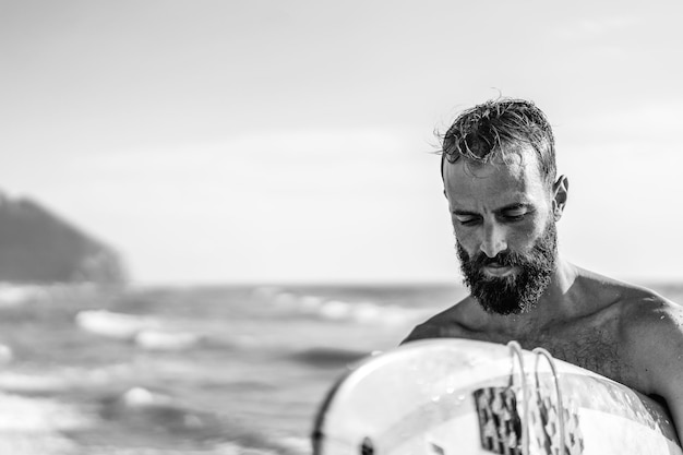 Surfer holding his surfboard walking on the beach hipster man\
training with surfboard lifestyle and freedom concept