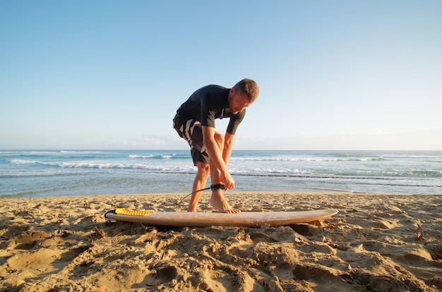 Surfer man fastens leash at leg, going to surf in ocean.