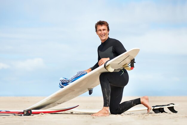 surfer man and portrait with surfboard on beach with wetsuit blue sky and cleaning with mock up space extreme sports athlete and person by ocean for training surfing workout and towel with smile