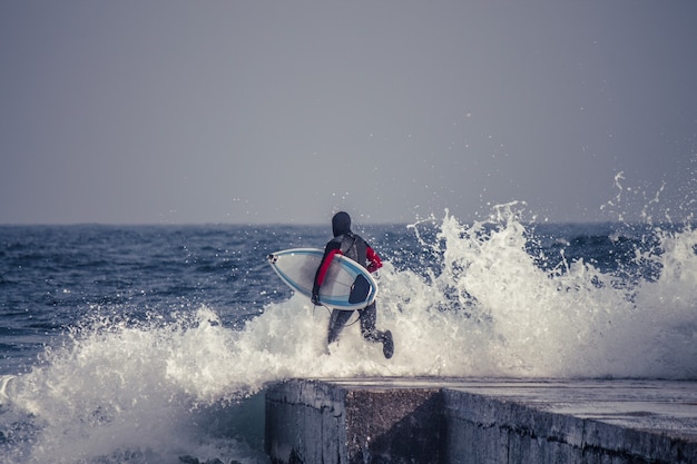 Surfer runs into water wearing a wetsuit in winter. Cold surfing. Wave splash.
