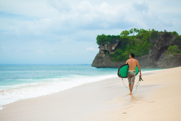 Surfer. uomo di surf con il surf che cammina sulla spiaggia tropicale di sabbia. stile di vita sano, attività acquatiche, sport acquatici. bellissimo oceano.