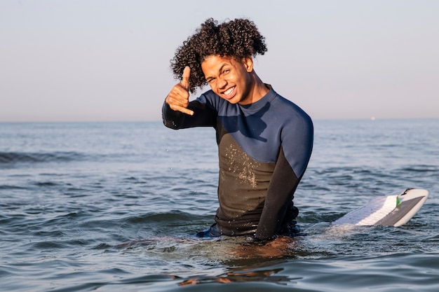 Surfer with afro hair sitting on his surfboard in the sea water sipping and doing the surfer salute with his hand.