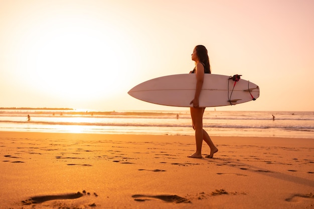 Una donna surfista su una spiaggia al tramonto con la tavola da surf che cammina verso il mare