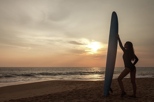 Surfer young woman on sandy beach with surf board watching sea waves at sunset