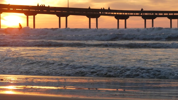 写真 カリフォルニアの日没時に桟橋の海の水波の人々と空でサーフィンするサーファー