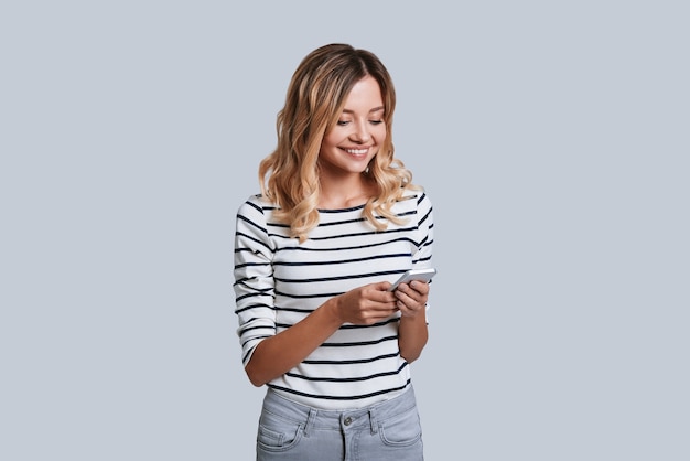 Surfing the net. Attractive young woman smiling and using her smart phone while standing against grey background