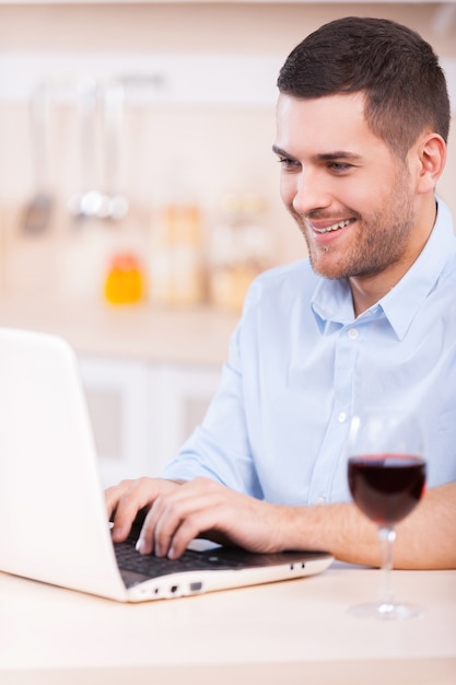 Navigare in rete in cucina. bel giovane in camicia che lavora al computer portatile mentre è seduto in cucina