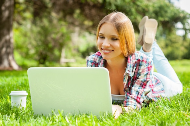 Surfing the net outdoors. beautiful young female student working on laptop and smiling while lying on the grass in park