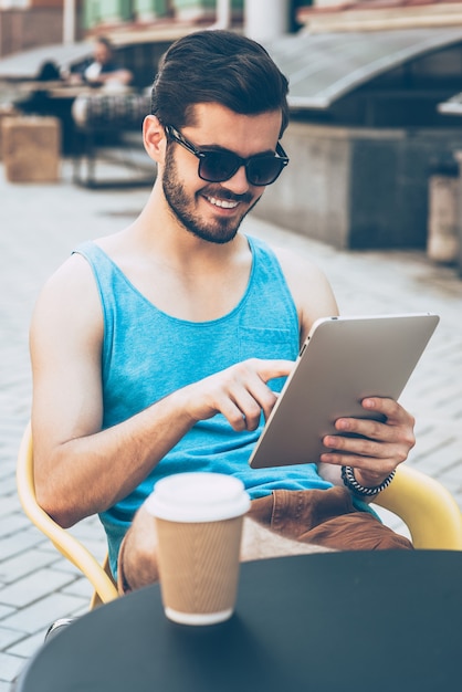 カフェからネットサーフィン。デジタルタブレットを持って、屋外の歩道のカフェに座って笑顔でそれを見ているカジュアルな服装のハンサムな若い男