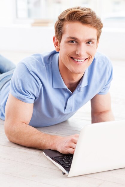 Surfing web at home. Handsome young man working on laptop and smiling while lying on the floor at his apartment