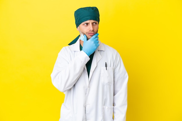 노란색 배경에 격리된 녹색 제복을 입은 외과의사 금발 남자는 의심을 갖고 얼굴 표정을 혼란스럽게 합니다.