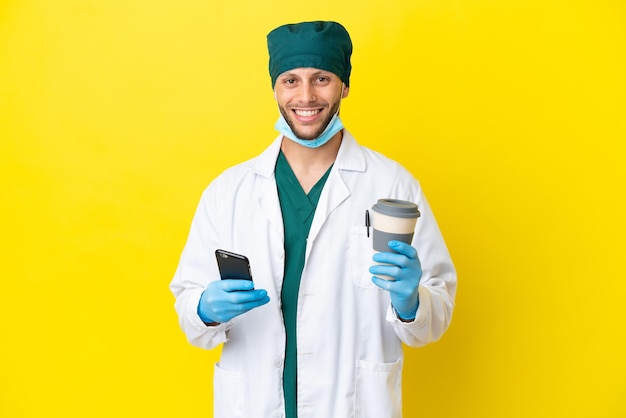 Chirurgo biondo in uniforme verde isolato su sfondo giallo che tiene il caffè da portare via e un cellulare