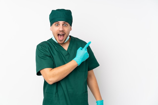 Surgeon in green uniform isolated on isolated white wall surprised and pointing side