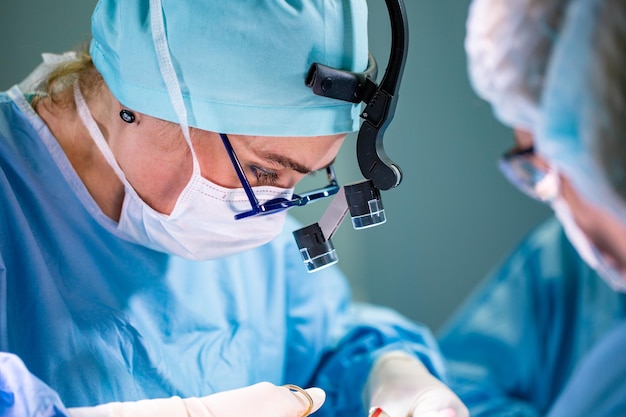 Surgeon and his assistant performing cosmetic surgery in hospital operating room Surgeon in mask wearing loupes during medical procadure Breast augmentation enlargement enhancement