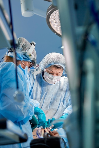 Surgeon and his assistant performing cosmetic surgery in hospital operating room Surgeon in mask wearing loupes during medical procadure Breast augmentation enlargement enhancement