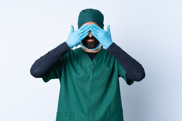 Surgeon man in green uniform over wall covering eyes by hands and smiling