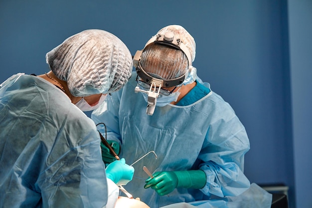 Surgeon performing cosmetic surgery in hospital operating room\
surgeon in mask wearing loupes during medical procadure breast\
augmentation enlargement enhancement blue background blue\
filter