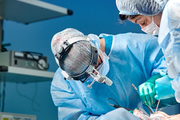 Surgeon performing cosmetic surgery in hospital operating room Surgeon in mask wearing loupes during medical procadure Breast augmentation enlargement enhancement Blue background blue filter