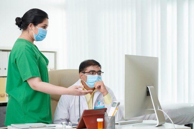 Surgeon and physician in protective masks discussing medical history of patient on computer screen