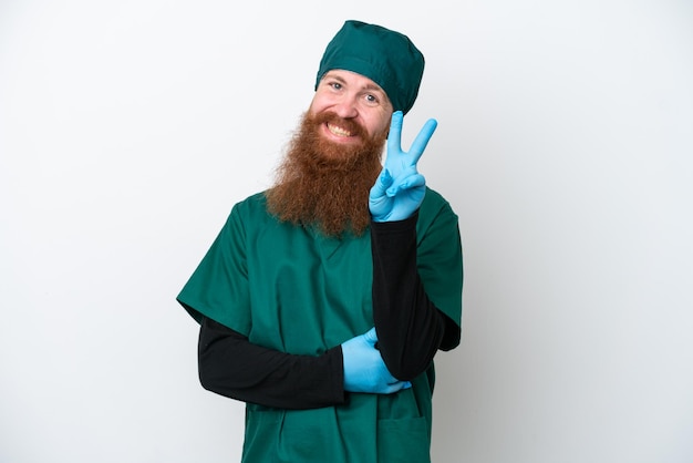 Surgeon redhead man in green uniform isolated on white background smiling and showing victory sign