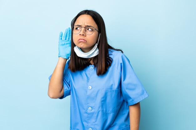 Surgeon woman over blue wall listening to something by putting hand on the ear