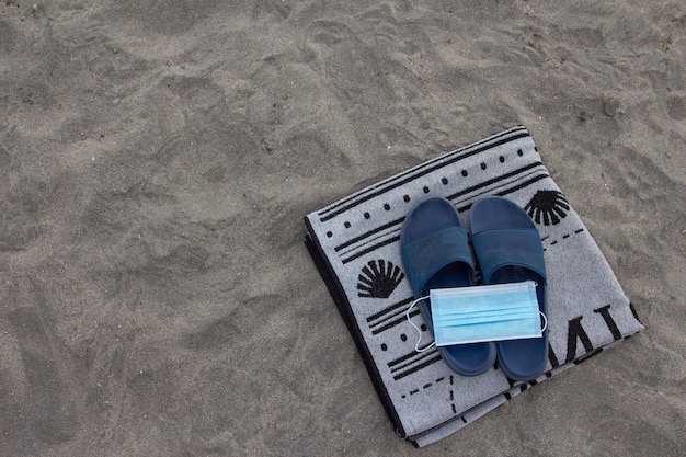 Surgery mask on top of flip flops and a towel on the beach.