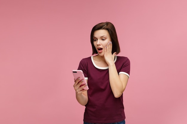 Surpised young brunette woman holding pink smartphone, smiling and expressing positivity.