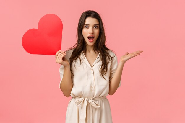 Surprise, relationship and love concept. Amused and wondered alluring woman didnt expact receive valentines day confession, open mouth gasping excited, holding red heart, pink wall