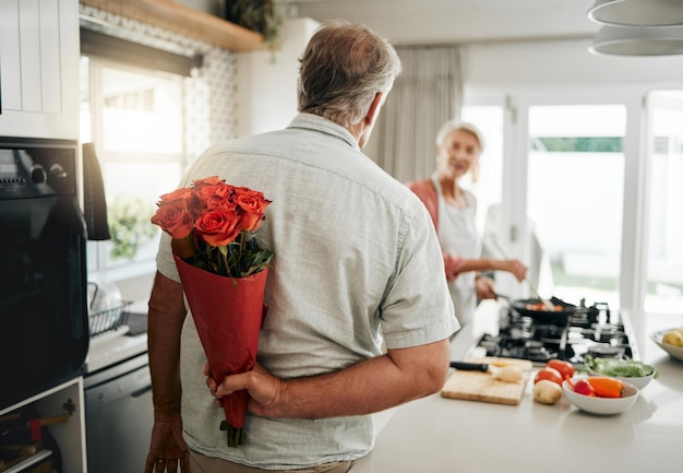 Surprise roses and a senior couple at home with romantic man\
spoiling wife with a bouquet of flowers on an anniversary birthday\
or valentines day at home husband hiding gift for a woman behind\
his