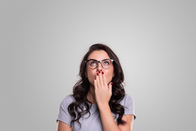 Surprised adult female in casual clothes and stylish eyeglasses covering mouth with hand and looking up with astonished face against gray background