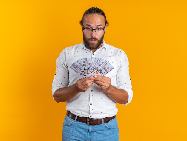 Surprised adult handsome man wearing glasses holding and looking at money isolated on orange wall with copy space