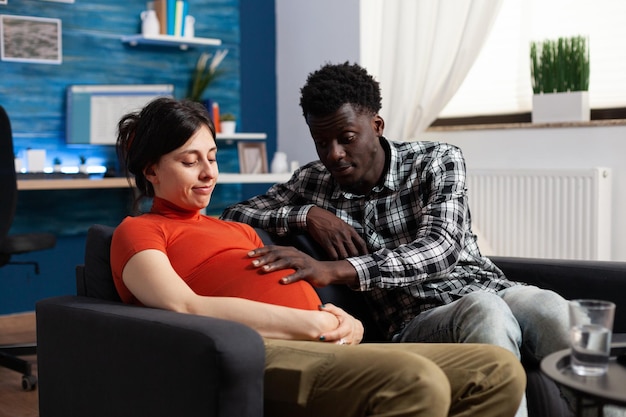 Surprised african american future father touching happy pregnant belly bump while sitting together in living room. Happy diverse adult couple expecting baby child while relaxing on sofa