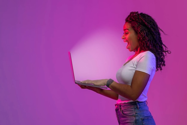 Удивленная афроамериканка смотрит на светящийся экран ноутбука, вид сбоку