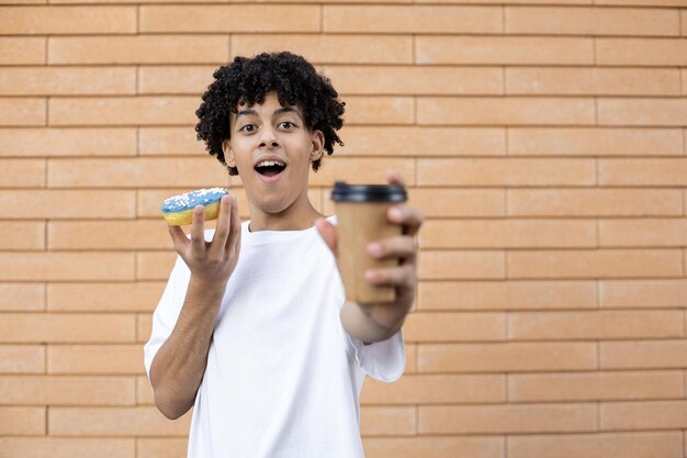Surprised AfricanAmerican guy holding a cup of coffee and a blue doughnut wearing a white tshirt