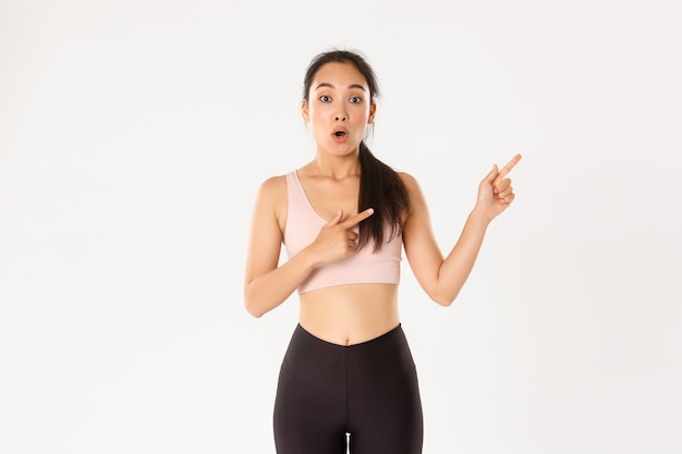 Surprised and amazed asian girl in fitness clothing, pointing fingers upper right corner, gasping and say wow impressed.
