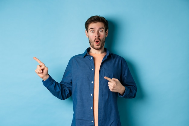 Surprised and amazed caucasian guy gasping in awe, pointing fingers left at logo, showing awesome promo, standing on blue background.