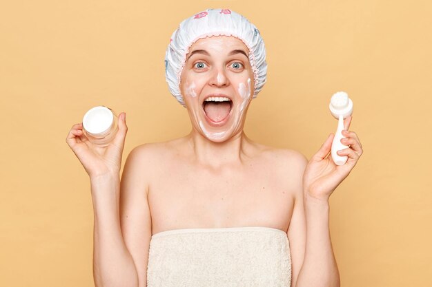 Surprised amazed excited woman wearing shower cap standing isolated over beige background holdingcleansing brush and cream for face skin care procedures