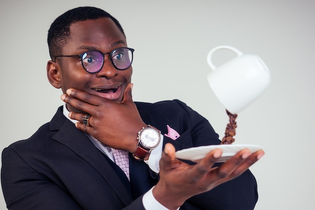 Surprised and amazed handsome afro american business man in a\
black classic suit and glasses holding a cup with flying coffee\
bean splash on plate in white background studio shot. magic morning\
drink