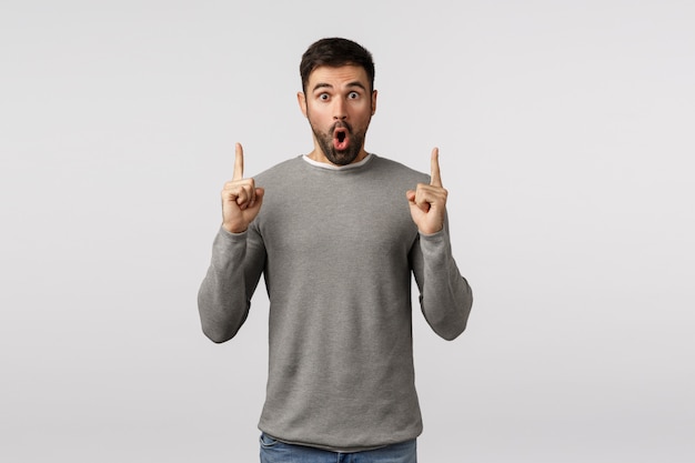 Surprised and amazed, speechless excited handsome bearded guy in grey sweater, say wow, folding lips amused, gasping astonished, pointing fingers up to turn attention, discuss impressive offer