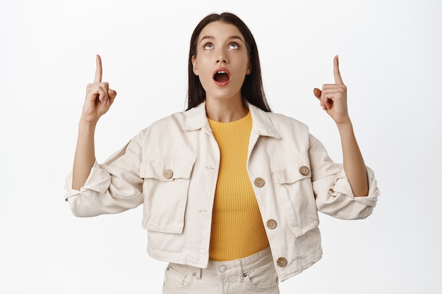 Surprised and amazed young woman pointing fingers, looking up with dropped jaw, wow sound, gasping fascinated, checking out sale banner, logo announcement on top on white.