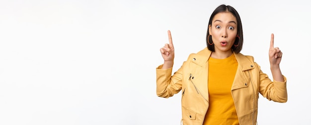 Surprised asian girl pointing fingers up express interest showing advertisement ahead demonstrating promo banner on top standing against white background