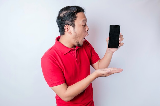 Surprised Asian man wearing red tshirt pointing at his smartphone isolated by white background