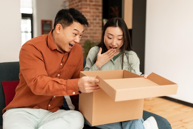 Surprised asian man and woman looking inside cardboard box satisfied with ordered item living room